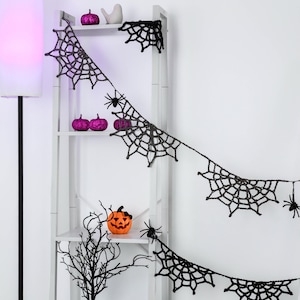 May include: A black crocheted spider web garland featuring multiple web segments and small black spider attachments, draped across a white shelving unit decorated with purple glitter pumpkins and a small orange jack-o'-lantern.