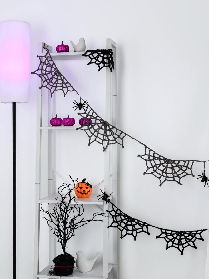 May include: A black crocheted spider web garland featuring multiple web segments and small black spider attachments, draped across a white shelving unit decorated with purple glitter pumpkins and a small orange jack-o'-lantern.