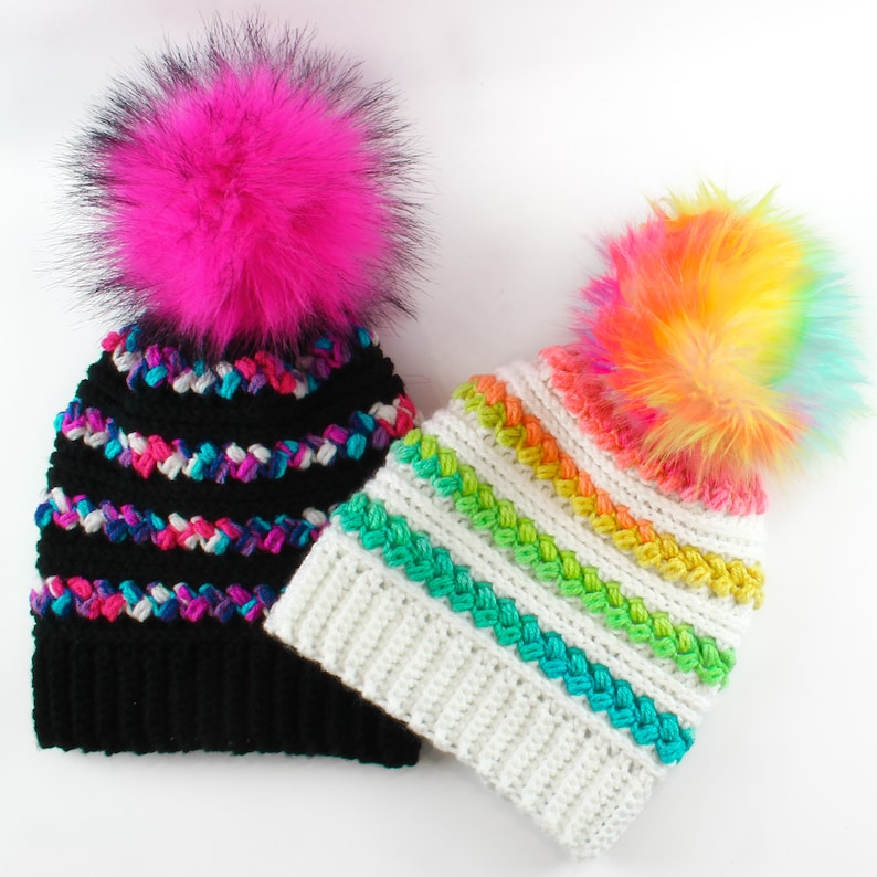 Puede incluir: Dos gorros de ganchillo con rayas de colores y pompones de piel sint&eacute;tica. Un gorro es negro con un pomp&oacute;n rosa, el otro es blanco con un pomp&oacute;n arco&iacute;ris.