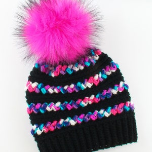 Puede incluir: Boina de crochet negra con un pomp&oacute;n de piel sint&eacute;tica rosa. La boina tiene rayas de colores rosa, azul, blanco y morado.