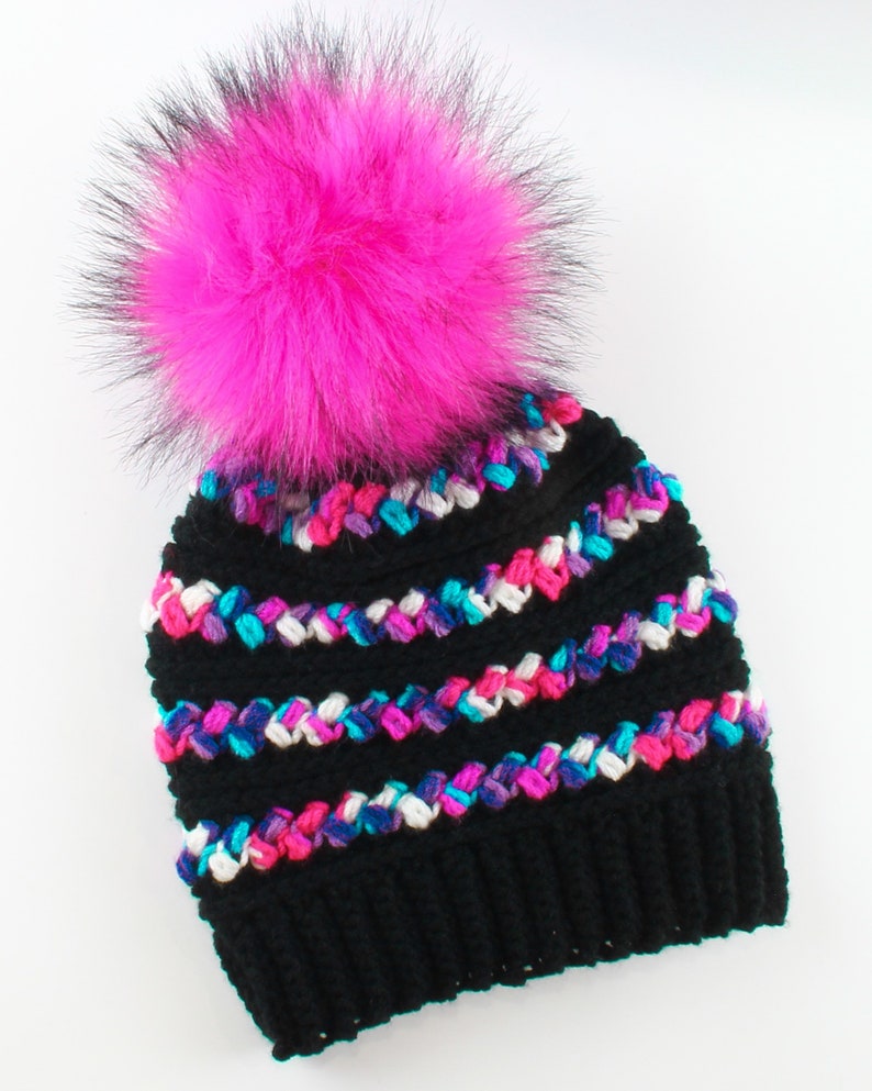 Puede incluir: Boina de crochet negra con un pomp&oacute;n de piel sint&eacute;tica rosa. La boina tiene rayas de colores rosa, azul, blanco y morado.