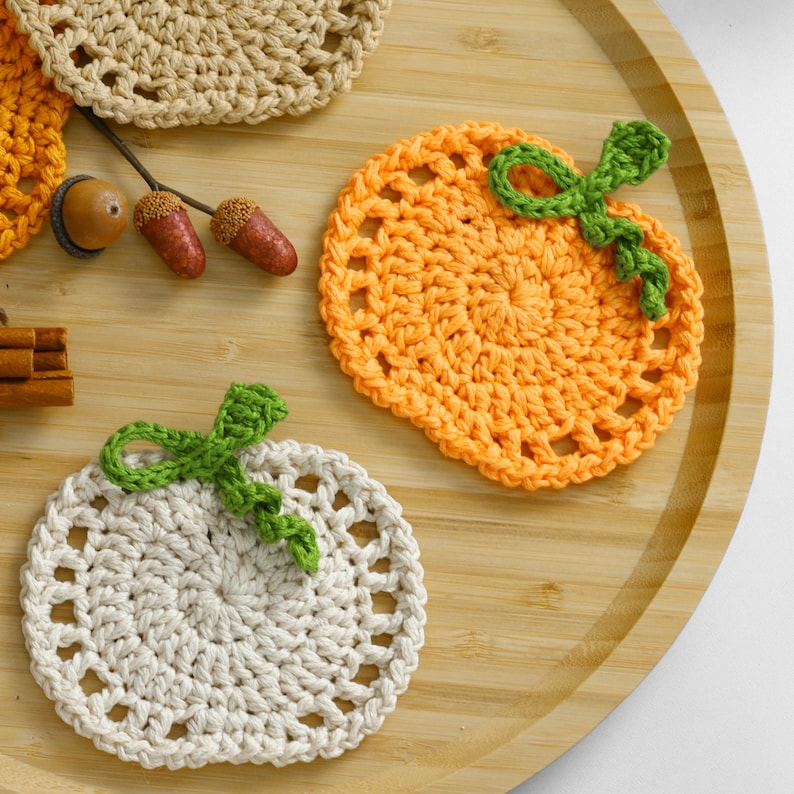 May include: Two round crochet coasters shaped like pumpkins, one in orange and one in off-white, each featuring a green crocheted stem and leaf. Also included are two small brown acorns and a partial view of a tan crochet piece.