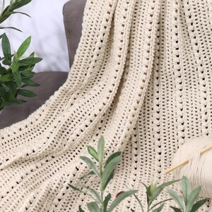 Pu&ograve; includere: Coperta all'uncinetto color crema drappeggiata su una sedia grigia. La coperta ha un design a trama aperta e strutturata. Un gomitolo di filo e ferri da maglia poggiano sulla coperta. Piante verdi e frondose in primo piano e sullo sfondo.
