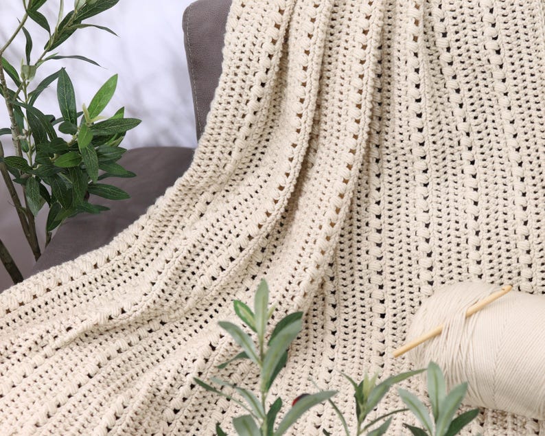 Pu&ograve; includere: Coperta all'uncinetto color crema drappeggiata su una sedia grigia. La coperta ha un design a trama aperta e strutturata. Un gomitolo di filo e ferri da maglia poggiano sulla coperta. Piante verdi e frondose in primo piano e sullo sfondo.