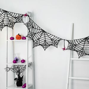 May include: A black crocheted spider web garland featuring intricate web patterns and small pink spider accents. The garland is draped across a white shelving unit and a white ladder, creating a decorative display for the holiday season.