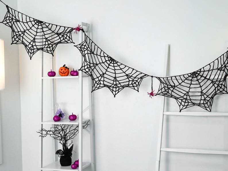 May include: A black crocheted spider web garland featuring intricate web patterns and small pink spider accents. The garland is draped across a white shelving unit and a white ladder, creating a decorative display for the holiday season.
