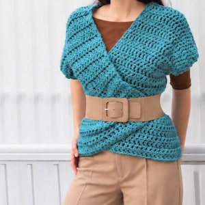 May include: A teal crochet wrap top with short sleeves and a V-neckline, layered over a brown shirt. The top is cinched at the waist with a wide beige elastic belt featuring a large square buckle, paired with tan trousers.