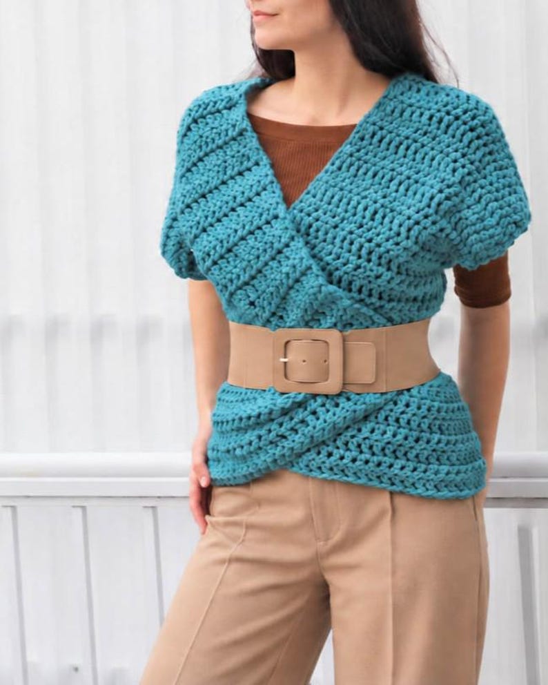 May include: A teal crochet wrap top with short sleeves and a V-neckline, layered over a brown shirt. The top is cinched at the waist with a wide beige elastic belt featuring a large square buckle, paired with tan trousers.