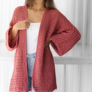 Pu&ograve; includere: Un cardigan all'uncinetto di colore rosa polvere con una vestibilit&agrave; ampia e maniche lunghe. Il cardigan &egrave; aperto sul davanti e presenta un semplice motivo a punto.