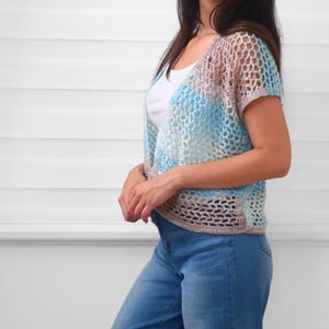 May include: A short-sleeved crochet cardigan featuring an open mesh stitch pattern in a variegated color palette of light blue, soft pink, and white. The garment has a V-neckline and a cropped length, worn over a plain white tank top and blue jeans.