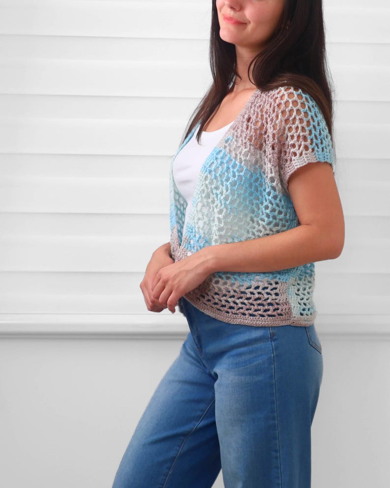 May include: A short-sleeved crochet cardigan featuring an open mesh stitch pattern in a variegated color palette of light blue, soft pink, and white. The garment has a V-neckline and a cropped length, worn over a plain white tank top and blue jeans.