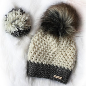 May include: A white and grey crocheted beanie with a faux fur pom pom. The beanie has a grey band at the bottom and a brown leather tag that says "The Easy Design".
