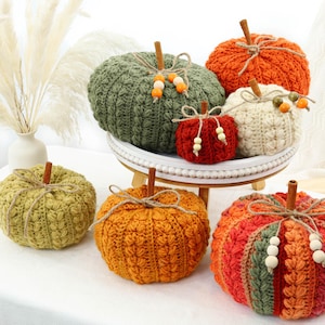 May include: A collection of seven crocheted pumpkins in various sizes and colors including olive green, bright orange, cream, and deep red. Each pumpkin features a cinnamon stick stem, twine bow, and wooden beads. Some pumpkins sit on a white tray.
