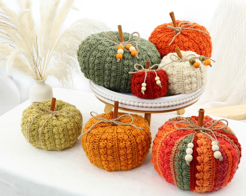 May include: A collection of seven crocheted pumpkins in various sizes and colors including olive green, bright orange, cream, and deep red. Each pumpkin features a cinnamon stick stem, twine bow, and wooden beads. Some pumpkins sit on a white tray.