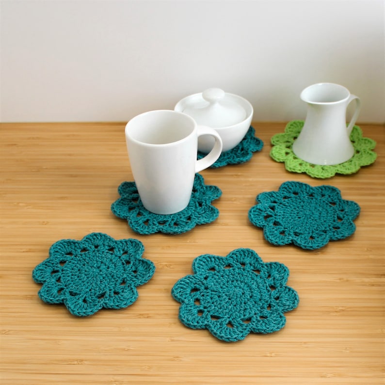 Op de afbeelding: Vijf gehaakte onderzetters in teal en groen. De onderzetters hebben de vorm van een bloem met meerdere lagen gehaakte steken. Twee van de onderzetters hebben witte keramische mokken en een suikerpot erop.