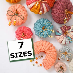 May include: A collection of crocheted pumpkins in various colors including peach, teal, pink, and mauve. Each pumpkin features a cinnamon stick stem, twine, and small wooden beads. A white square sign with the text 7 SIZES is visible.