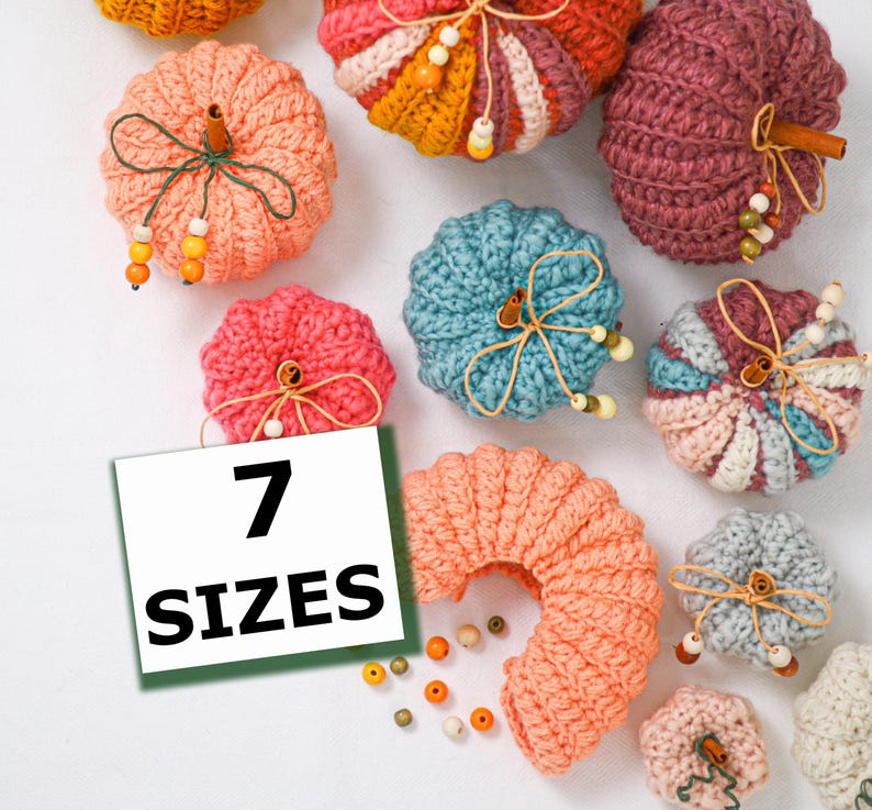 May include: A collection of crocheted pumpkins in various colors including peach, teal, pink, and mauve. Each pumpkin features a cinnamon stick stem, twine, and small wooden beads. A white square sign with the text 7 SIZES is visible.