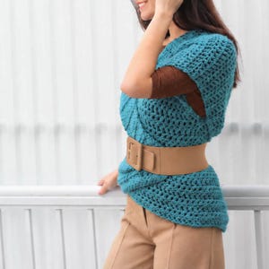 May include: A teal blue sleeveless crochet top featuring a textured knit pattern, layered over a brown long-sleeve shirt. The top is cinched at the waist with a wide, smooth tan belt that has a rectangular buckle, paired with tan trousers.
