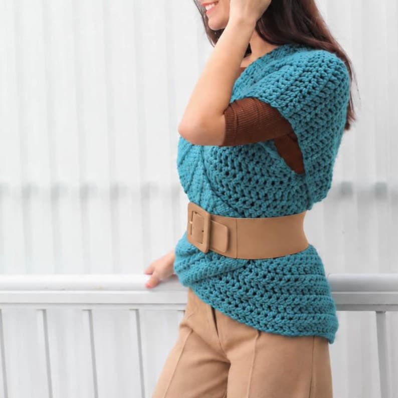 May include: A teal blue sleeveless crochet top featuring a textured knit pattern, layered over a brown long-sleeve shirt. The top is cinched at the waist with a wide, smooth tan belt that has a rectangular buckle, paired with tan trousers.