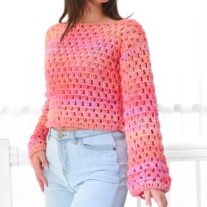 May include: A cropped, long-sleeved crochet sweater in shades of pink and orange. The open weave design reveals the skin beneath. The sweater has a wide neckline and is paired with light blue jeans.