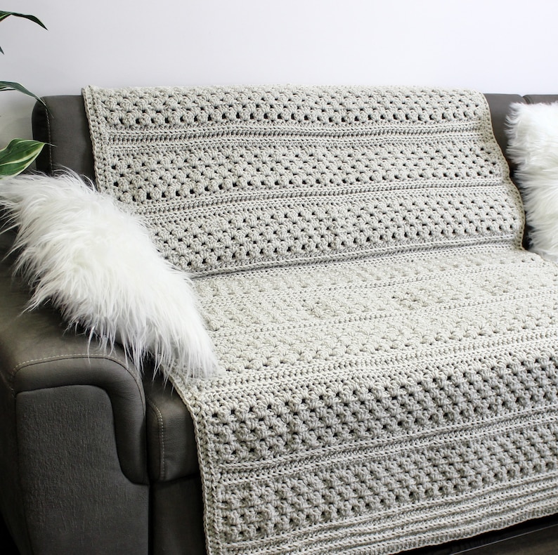 May include: A light gray crocheted throw blanket draped over a gray couch with a white faux fur pillow. The blanket has a textured stitch pattern.