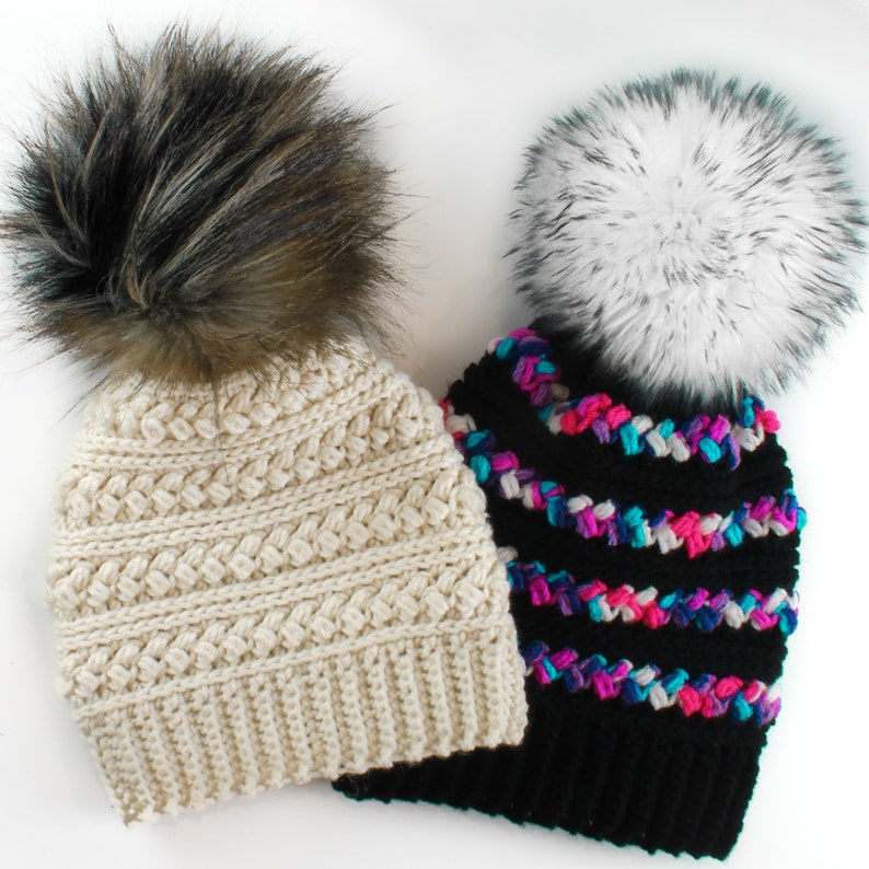Puede incluir: Dos gorros de punto con pompones de piel sint&eacute;tica. Un gorro es crema con un pomp&oacute;n marr&oacute;n y el otro es negro con un pomp&oacute;n blanco y rayas de colores.