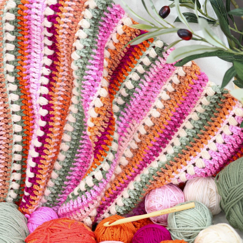 Pu&ograve; includere: Una colorata coperta all'uncinetto fatta a mano nei toni del rosa, arancione, verde e crema. La coperta presenta un motivo strutturato ed &egrave; circondata da gomitoli di lana e un uncinetto, suggerendo un progetto artigianale.