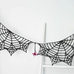 May include: A black crocheted spider web garland featuring several small, bright pink plastic spiders attached to the web structure. The garland is draped horizontally, showcasing intricate knotted yarn patterns and pointed edges.