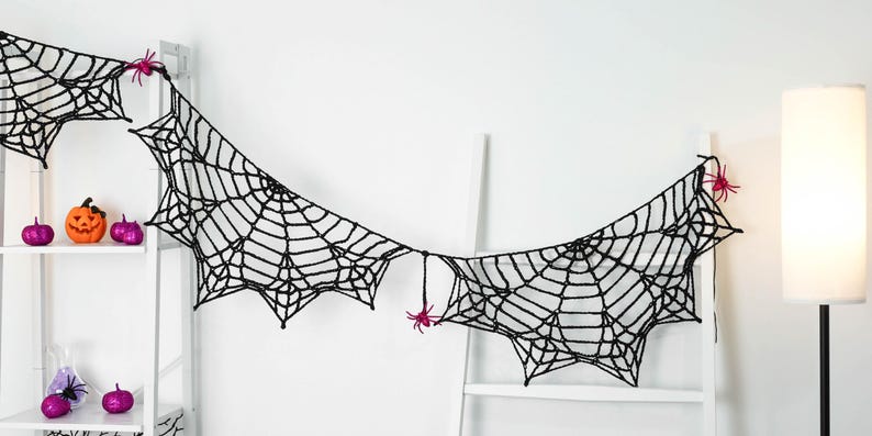 May include: A black crocheted spider web garland featuring several small, bright pink plastic spiders attached to the web structure. The garland is draped horizontally, showcasing intricate knotted yarn patterns and pointed edges.