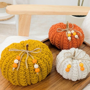 May include: Three crocheted pumpkins in shades of yellow, orange, and white, arranged on a wooden tray. Each pumpkin has a twine stem and is adorned with wooden beads. The pumpkins are a decorative item for the fall season.