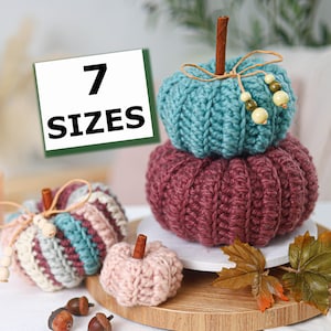 Peut inclure: Une collection de citrouilles au crochet en bleu sarcelle, bordeaux et rose clair, avec des tiges en bâton de cannelle et des perles en bois. Un panneau blanc avec le texte noir 7 SIZES. Les citrouilles sont sur une base ronde en bois.