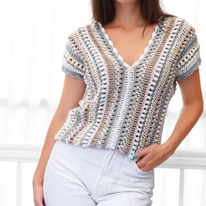 Puede incluir: Un top de crochet con cuello en V y mangas cortas, en una paleta de colores neutros de blanco, gris y marr&oacute;n. El top presenta un borde festoneado a lo largo del escote y las mangas. El patr&oacute;n de crochet crea un dise&ntilde;o de tejido abierto y texturizado.