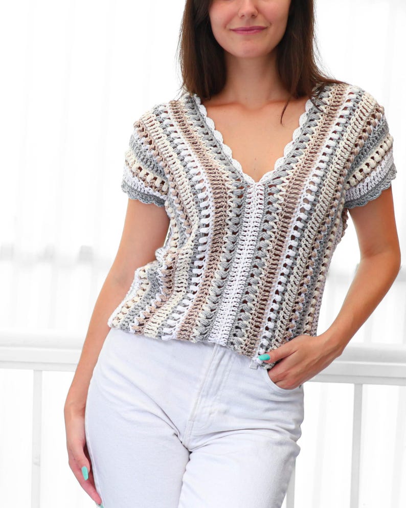 Puede incluir: Un top de crochet con cuello en V y mangas cortas, en una paleta de colores neutros de blanco, gris y marr&oacute;n. El top presenta un borde festoneado a lo largo del escote y las mangas. El patr&oacute;n de crochet crea un dise&ntilde;o de tejido abierto y texturizado.