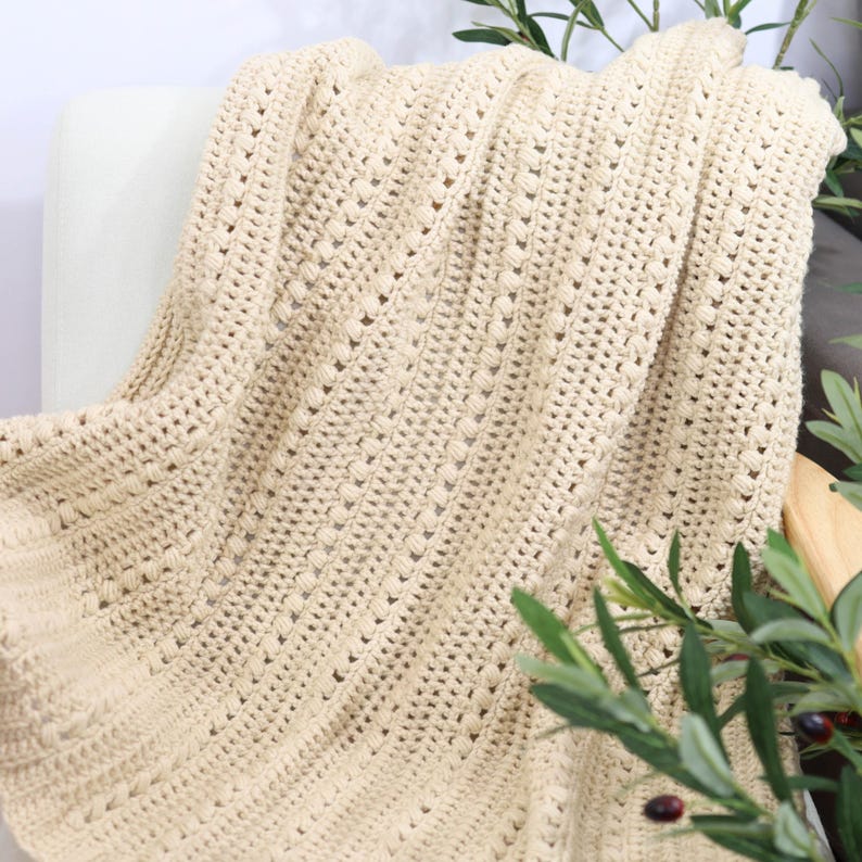 Pu&ograve; includere: Una coperta beige all'uncinetto drappeggiata su una sedia bianca. La coperta presenta un motivo strutturato con linee verticali e piccoli dettagli in rilievo. Il colore morbido e neutro e il design intricato suggeriscono un oggetto fatto a mano e accogliente.