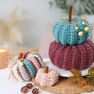May include: A collection of five crocheted pumpkins in teal, burgundy, and light pink. The pumpkins feature ribbed textures, cinnamon stick stems, and twine bows with wooden beads. One pumpkin is striped with teal, burgundy, and cream yarn.