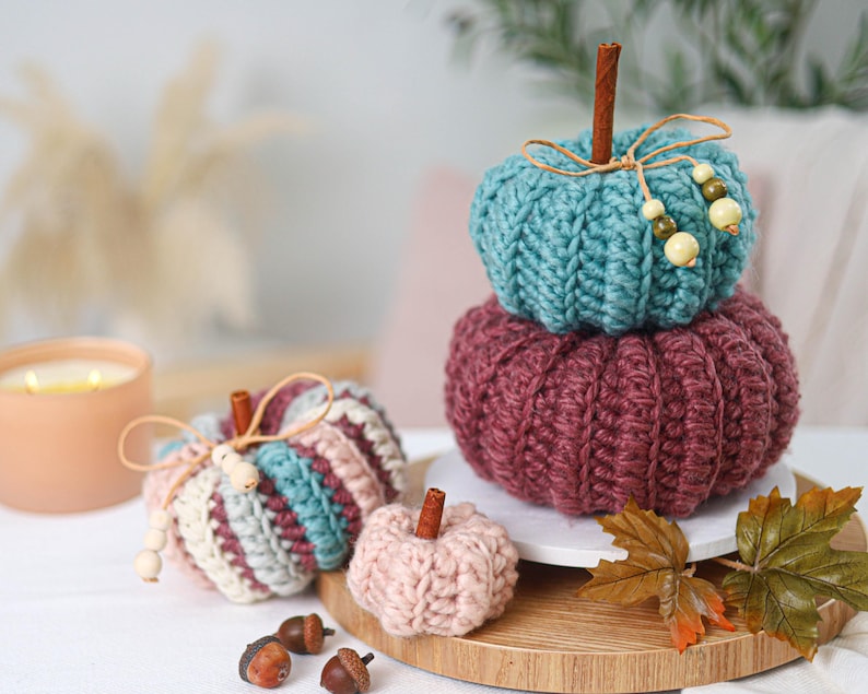 May include: A collection of five crocheted pumpkins in teal, burgundy, and light pink. The pumpkins feature ribbed textures, cinnamon stick stems, and twine bows with wooden beads. One pumpkin is striped with teal, burgundy, and cream yarn.