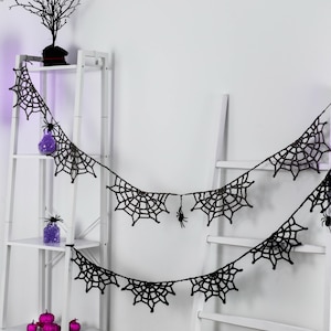 May include: A black crocheted garland featuring multiple spider web motifs connected by a thin string. Small black spider figures are attached to the web segments, creating a decorative hanging piece for seasonal display.