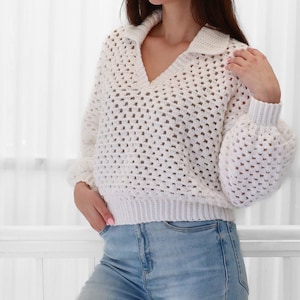 Peut inclure: Un pull en crochet blanc avec un col en V et un col montant. Le pull est ample et de longueur courte. Le pull est r&eacute;alis&eacute; avec un motif de point ouvert d&eacute;licat.