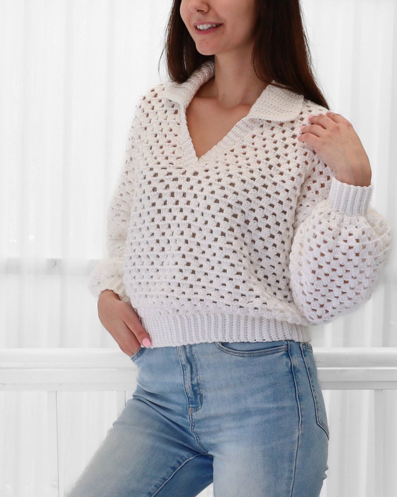 Peut inclure: Un pull en crochet blanc avec un col en V et un col montant. Le pull est ample et de longueur courte. Le pull est r&eacute;alis&eacute; avec un motif de point ouvert d&eacute;licat.