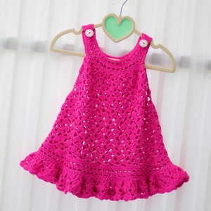 May include: A pink crocheted baby dress with a ruffled hem and white buttons. The dress is hanging on a wooden hanger with a heart-shaped cutout.