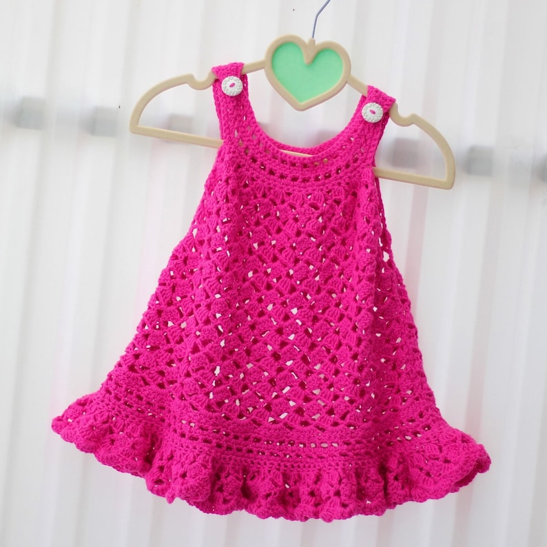 May include: A pink crocheted baby dress with a ruffled hem and white buttons. The dress is hanging on a wooden hanger with a heart-shaped cutout.