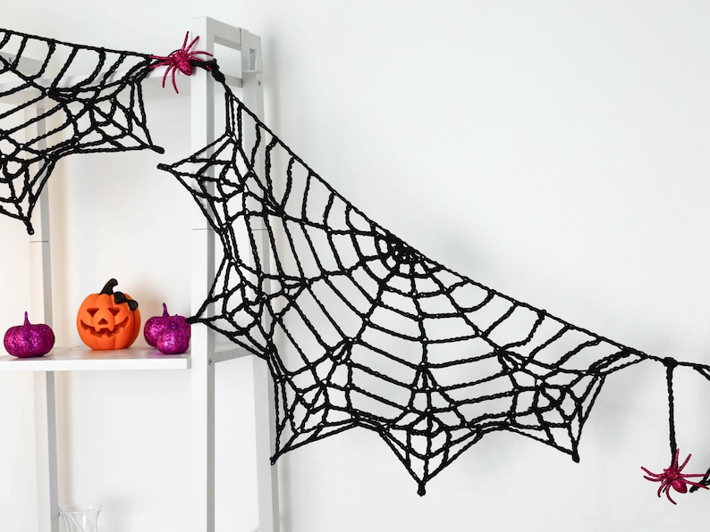 May include: A black crocheted spider web garland featuring intricate web patterns and small, bright pink plastic spiders attached at the connection points, designed for hanging as a decorative Halloween display.