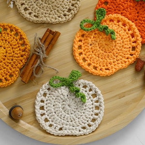 May include: A collection of round, crocheted pumpkin-shaped coasters in cream, light orange, and dark orange yarn. Each coaster features a green crocheted stem and leaf detail. A bundle of three cinnamon sticks tied with twine is included.