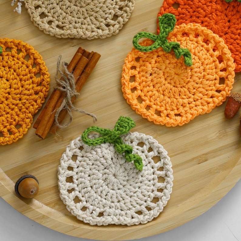 May include: A collection of round, crocheted pumpkin-shaped coasters in cream, light orange, and dark orange yarn. Each coaster features a green crocheted stem and leaf detail. A bundle of three cinnamon sticks tied with twine is included.