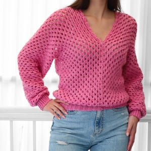 Pode incluir: Um su&eacute;ter de croch&ecirc; rosa com decote em V e mangas compridas. O su&eacute;ter tem um caimento solto e um delicado padr&atilde;o de ponto aberto.