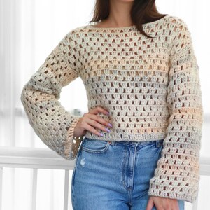 May include: A cream-colored crochet top with long sleeves and a square neckline. The open weave design features a gradient of colors, transitioning from cream to light peach and blue. The top is paired with blue jeans.