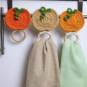 May include: Three crochet pumpkin toppers attached to wooden rings. Two pumpkins are orange and one is tan, each with a green vine. Two of the rings hold hanging kitchen towels, one with a tan gingham pattern and one in solid light green.