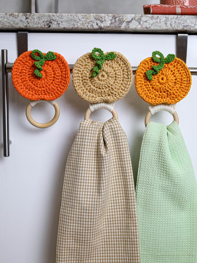 May include: Three crochet pumpkin toppers attached to wooden rings. Two pumpkins are orange and one is tan, each with a green vine. Two of the rings hold hanging kitchen towels, one with a tan gingham pattern and one in solid light green.