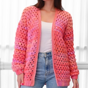 May include: A handmade, open-front crochet cardigan in shades of pink and coral. The long-sleeved sweater features a textured, open weave design. The cardigan is worn over a white top and blue jeans.