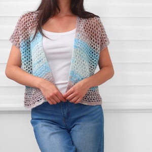 May include: A short-sleeved, open-front crochet cardigan featuring a loose, net-like stitch pattern. The garment displays a variegated color palette of soft brown, light blue, and white tones, layered over a plain white tank top and blue jeans.